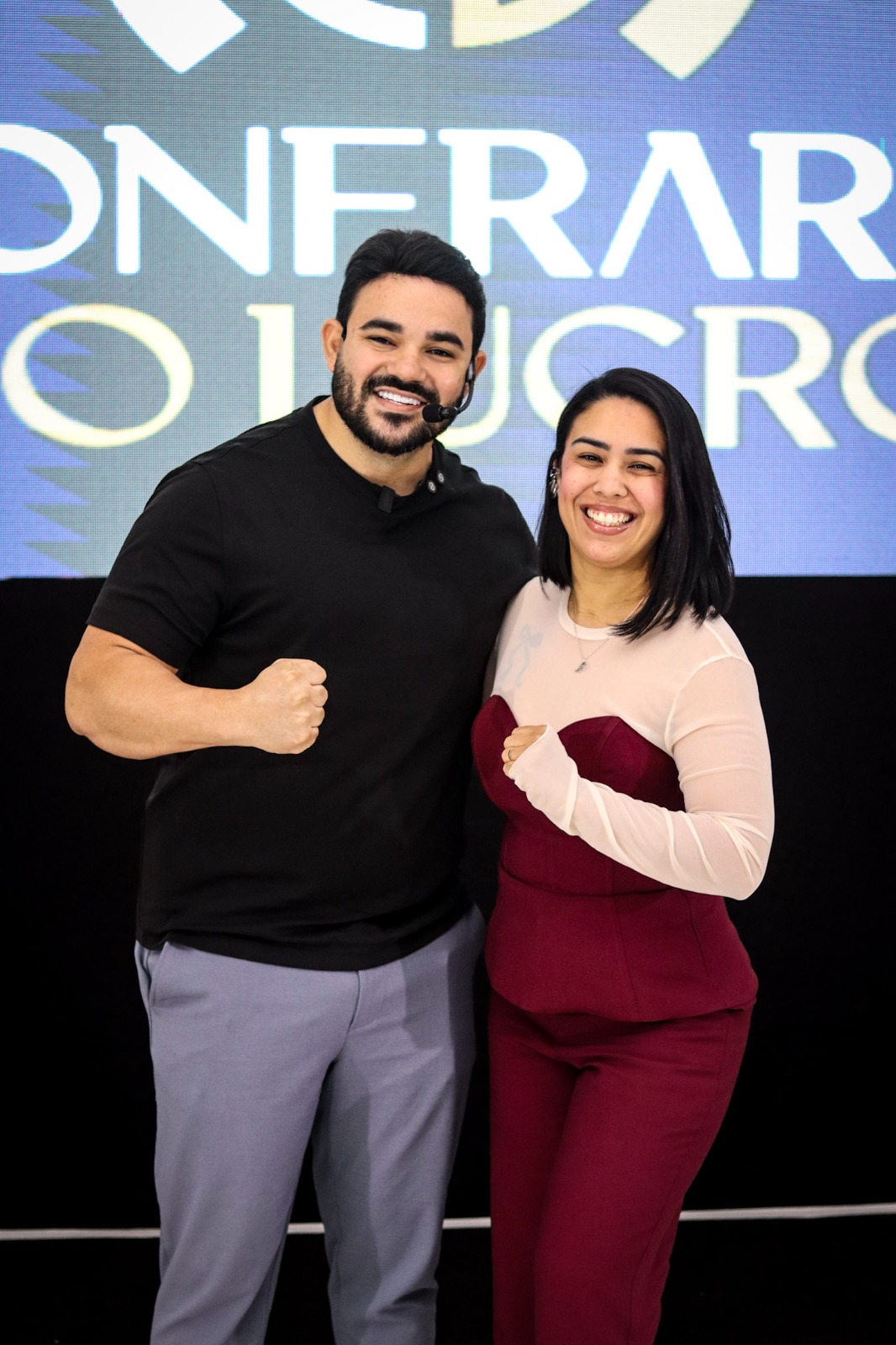 Diego e Ediana diante do telão da Confraria do Lucro, sorrindo com os punhos cerrados
